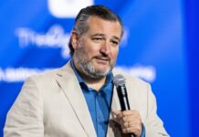 Man in beige blazer holding microphone with blue background.