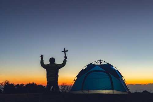 Person holding cross beside a tent at sunset.