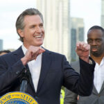 Newsom Declares 100% TAX WAR on Trump Funds… Smiling man giving a speech at an outdoor event.