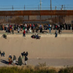 Drainage Tunnel BUST: 19 Captured in Shocking Border RAID…. Large group of people gathered along a tall border fence near a concrete channel