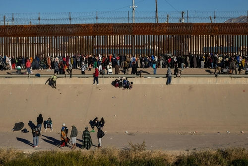 shutterstock2257563879jpg Large group of people gathered along a tall border fence near a concrete channel