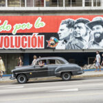 Spy Chief Lands in Cuba—Delivers Shocking Demand… A mural depicting revolutionary figures with a classic car passing by