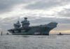 Stealth Mini-Carriers Quietly Rewrite War Aircraft carrier with tugboats on a cloudy sea.