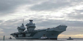 Aircraft carrier with tugboats on a cloudy sea.