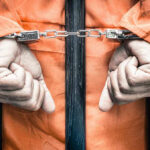 Handcuffed person in orange jumpsuit behind bars.