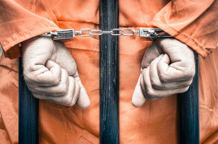 217094626 Handcuffed person in orange jumpsuit behind bars.