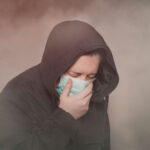 See The World’s Dirtiest Air Person with hood and mask coughing in foggy setting.