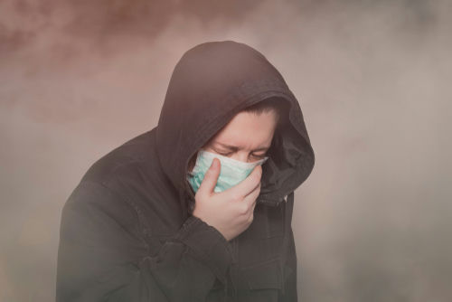 shutterstock_1610371663 Person with hood and mask coughing in foggy setting.