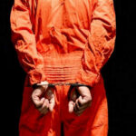 Repeat Offender Ends Up in Governor’s Backyard Person in orange jumpsuit handcuffed behind their back.