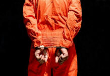 Repeat Offender Ends Up in Governor’s Backyard Person in orange jumpsuit handcuffed behind their back.