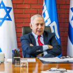 Netanyahu Drops BOMBSHELL About Iran’s ‘Hit List’ man in suit seated at desk with Israeli flags