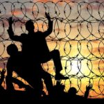 Silhouettes of people climbing a barbed wire fence at sunset