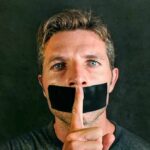 man with tape over mouth holding a finger to lips