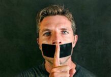 man with tape over mouth holding a finger to lips