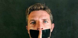 man with tape over mouth holding a finger to lips