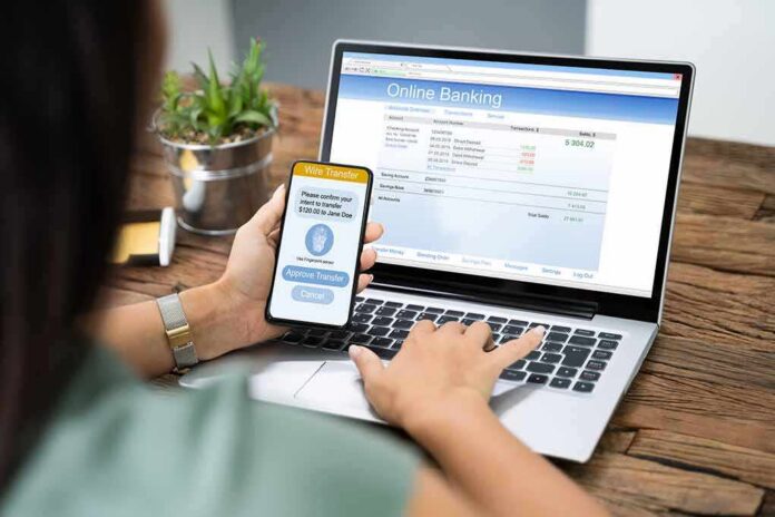 shutterstock_1542438851 Person using smartphone to approve bank transfer beside laptop with online banking