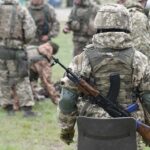 Soldier in camouflage with rifle and body armor stands near a group in a field