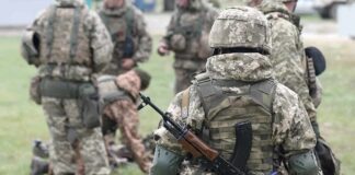 Soldier in camouflage with rifle and body armor stands near a group in a field