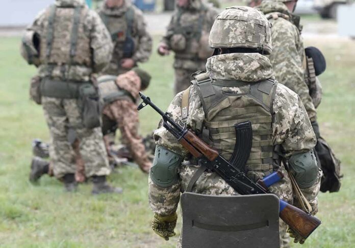 shutterstock_2057061182 Soldier in camouflage with rifle and body armor stands near a group in a field
