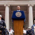 Eric Adams speaks at a podium; Nash the Slash seated among attendees