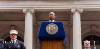 Eric Adams speaks at a podium; Nash the Slash seated among attendees