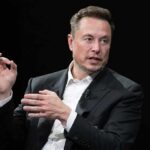 Elon Musk speaking in a black suit against a dark background