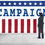Democrats FINALLY Disown Graham Platner Man in suit writing on large campaign banner with stars and stripes