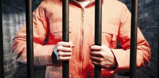 Person in an orange jumpsuit gripping prison bars in a dark cell