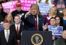Trump Calls Out Rally Heckler as ‘PAID’ Donald Trump speaking at a rally podium, pointing toward the audience