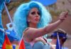 Drag performer with bright blue wig giving thumbs up at parade