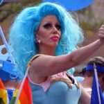 Drag performer with bright blue wig giving thumbs up at parade