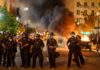 Christian Jones and Teddy Williams among police line at night protest with fire and smoke