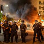 52 Cops HURT During ICE Protest While Mayoral Candidate Blames Police Christian Jones and Teddy Williams among police line at night protest with fire and smoke