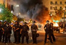 Christian Jones and Teddy Williams among police line at night protest with fire and smoke