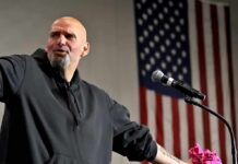 Fetterman Warns Democrats of a SOCIALISM Problem Bald man speaking at a microphone with a U.S. flag in the background